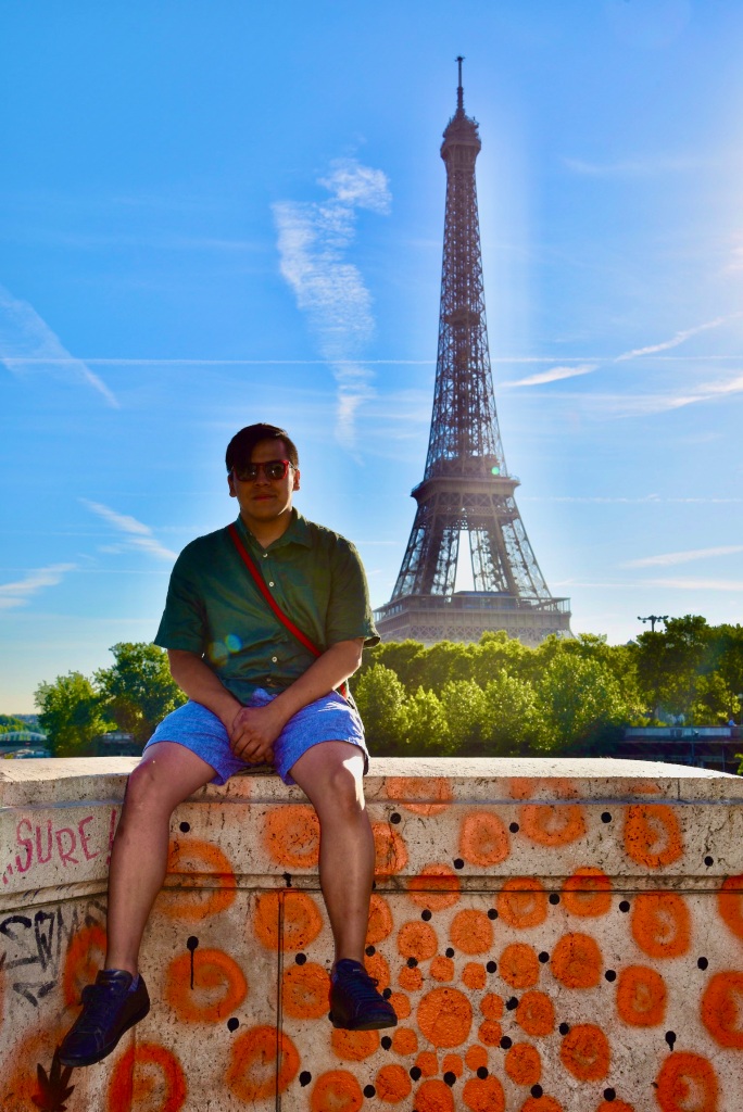 A photo of Emmanuel Leal in Paries, sitting on a graffitied ledge with the Eiffel Tower surrounded by trees in the background.